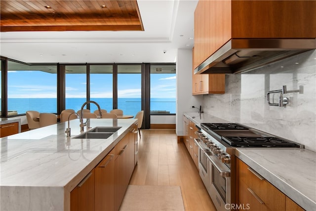 kitchen with range with two ovens, extractor fan, a sink, light stone countertops, and a large island with sink