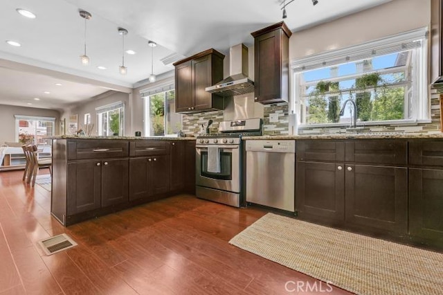 kitchen featuring dark wood-style floors, a peninsula, dark brown cabinets, appliances with stainless steel finishes, and wall chimney exhaust hood