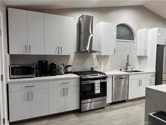 kitchen featuring appliances with stainless steel finishes, vaulted ceiling, white cabinets, sink, and wall chimney range hood