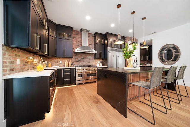 kitchen with high end appliances, wall chimney range hood, pendant lighting, a kitchen island, and light wood-type flooring