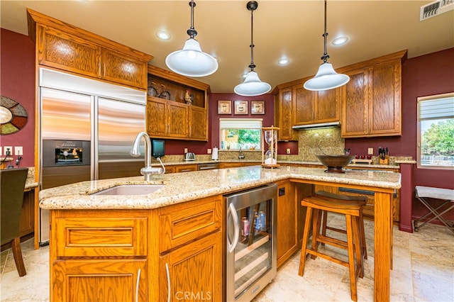 kitchen with sink, a kitchen island with sink, light stone counters, decorative light fixtures, and beverage cooler