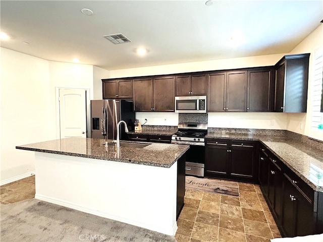 kitchen featuring dark stone countertops, stainless steel appliances, dark brown cabinets, a kitchen island with sink, and sink