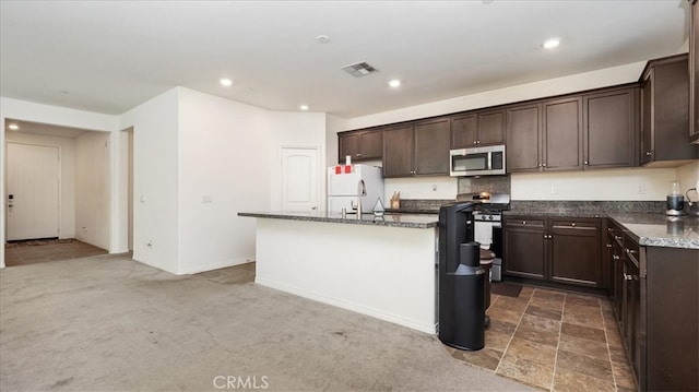 kitchen featuring appliances with stainless steel finishes, dark stone countertops, an island with sink, dark brown cabinets, and dark carpet