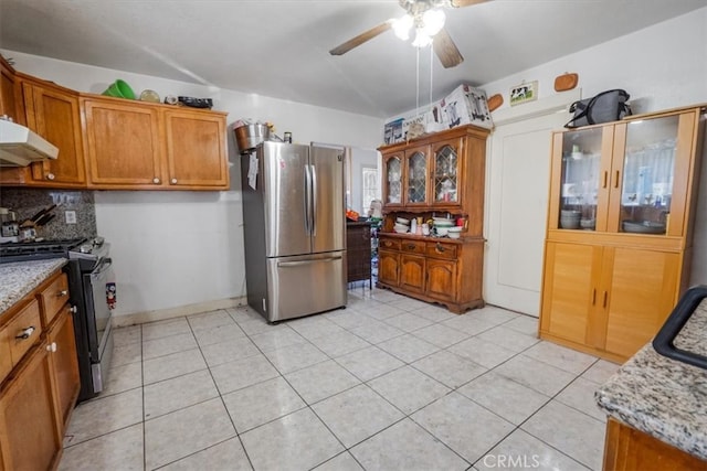 kitchen with brown cabinetry, decorative backsplash, appliances with stainless steel finishes, light stone countertops, and under cabinet range hood