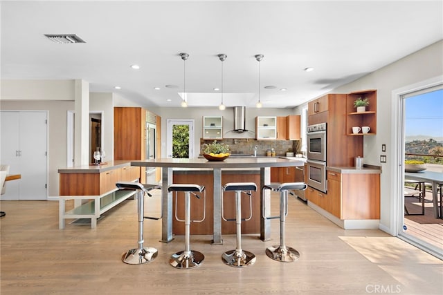 kitchen featuring wall chimney range hood, light wood-type flooring, stainless steel double oven, and hanging light fixtures