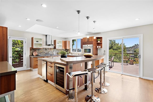 kitchen with wall chimney range hood, light hardwood / wood-style flooring, an island with sink, pendant lighting, and beverage cooler