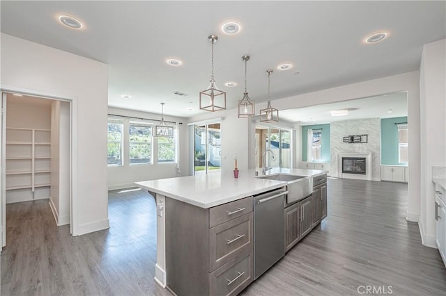 kitchen featuring stainless steel dishwasher, a large fireplace, a kitchen island with sink, hardwood / wood-style floors, and hanging light fixtures