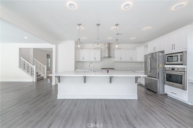 kitchen featuring wall chimney exhaust hood, stainless steel appliances, wood-type flooring, a center island with sink, and white cabinets