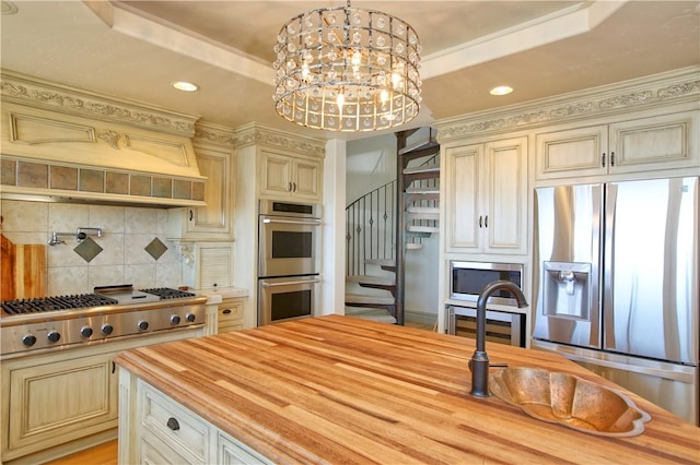kitchen with a tray ceiling, stainless steel appliances, wooden counters, cream cabinets, and backsplash