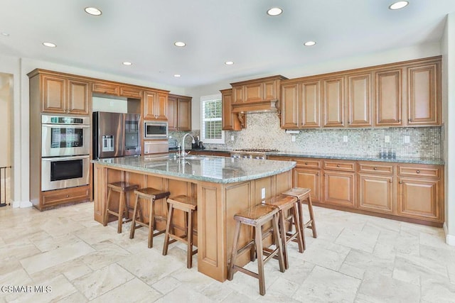 kitchen with a kitchen breakfast bar, light stone counters, custom exhaust hood, stainless steel appliances, and an island with sink