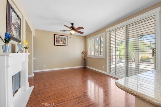 living room with a glass covered fireplace, wood finished floors, a ceiling fan, and baseboards