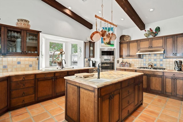 kitchen featuring tile counters, stainless steel appliances, a center island with sink, lofted ceiling with beams, and tasteful backsplash