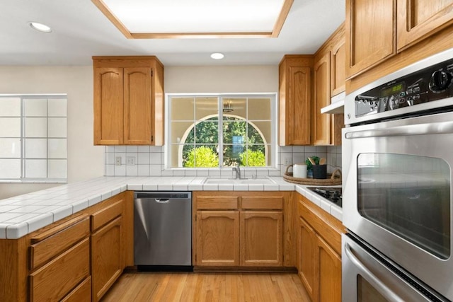 kitchen with appliances with stainless steel finishes, tile countertops, light wood-type flooring, and tasteful backsplash