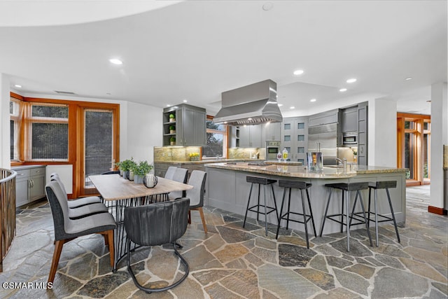 kitchen with island exhaust hood, a kitchen island with sink, built in appliances, gray cabinets, and light stone counters