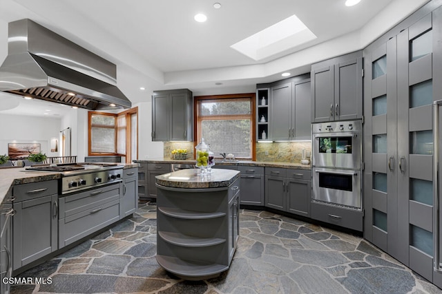 kitchen featuring a skylight, backsplash, stainless steel appliances, gray cabinets, and range hood