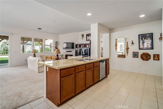 kitchen featuring a center island with sink, sink, stainless steel dishwasher, light colored carpet, and a tiled fireplace