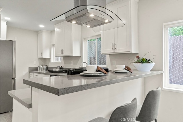 kitchen with stainless steel refrigerator, kitchen peninsula, island range hood, white cabinetry, and a kitchen bar