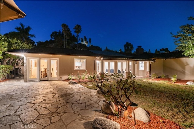 back of house at night featuring stucco siding, a patio area, a chimney, and french doors