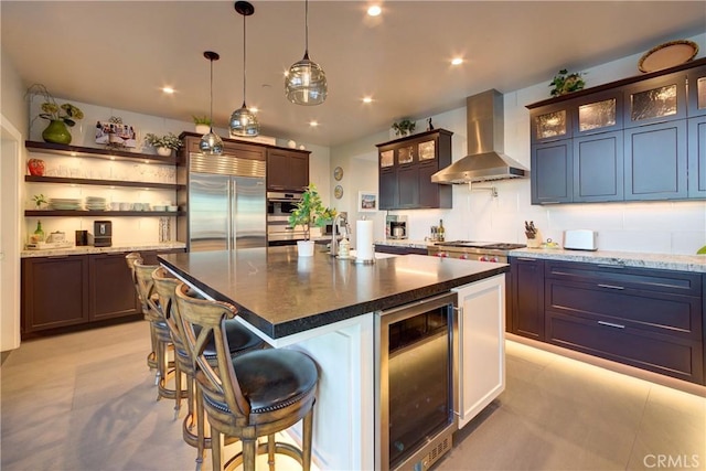 kitchen featuring wine cooler, stainless steel built in refrigerator, dark brown cabinets, an island with sink, and wall chimney range hood