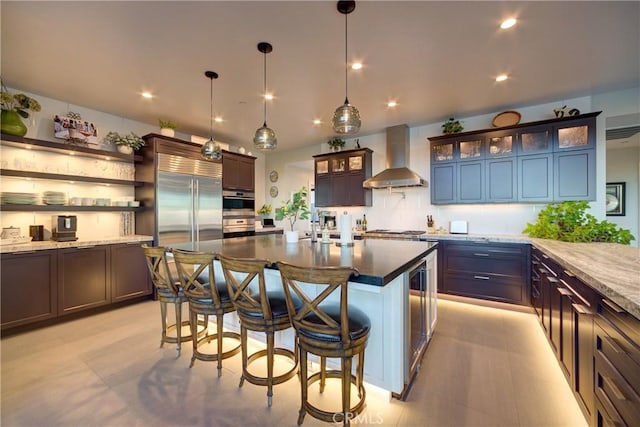 kitchen with decorative light fixtures, dark stone counters, stainless steel appliances, a kitchen island with sink, and wall chimney range hood