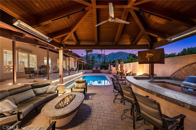 pool at dusk featuring a patio area, an outdoor living space with a fire pit, a gazebo, ceiling fan, and an outdoor bar