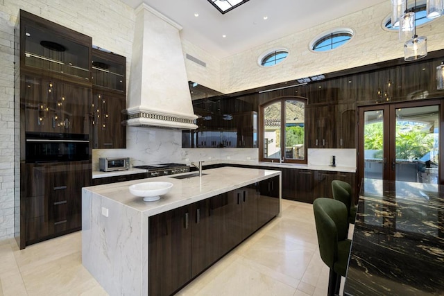 kitchen with premium range hood, a center island with sink, oven, decorative backsplash, and stainless steel gas cooktop