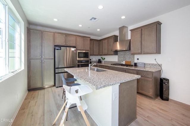 kitchen with plenty of natural light, wall chimney range hood, stainless steel appliances, and an island with sink