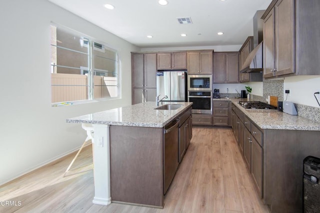 kitchen with light stone countertops, stainless steel appliances, wall chimney range hood, a kitchen island with sink, and light wood-type flooring