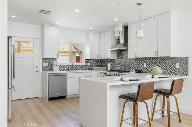 kitchen with appliances with stainless steel finishes, white cabinetry, wall chimney exhaust hood, kitchen peninsula, and light hardwood / wood-style flooring