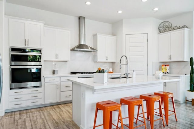 kitchen featuring a breakfast bar, a kitchen island with sink, sink, wall chimney range hood, and white cabinetry