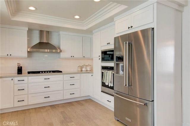 kitchen featuring appliances with stainless steel finishes, white cabinets, light wood-type flooring, a raised ceiling, and wall chimney exhaust hood