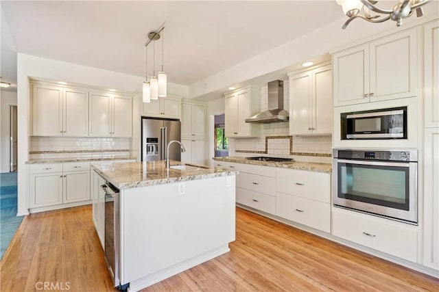 kitchen with white cabinets, wall chimney range hood, light wood-type flooring, an island with sink, and appliances with stainless steel finishes