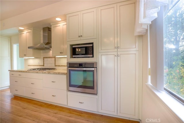 kitchen featuring wall chimney range hood, light hardwood / wood-style flooring, decorative backsplash, white cabinetry, and stainless steel appliances