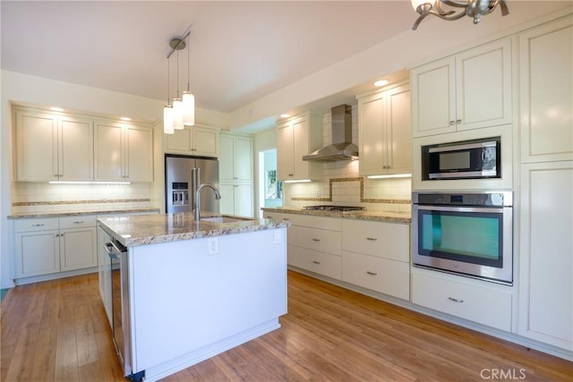 kitchen featuring sink, stainless steel appliances, wall chimney range hood, light hardwood / wood-style floors, and a center island with sink