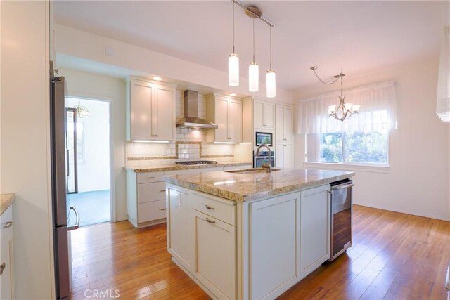 kitchen with sink, wall chimney range hood, decorative light fixtures, a center island with sink, and light hardwood / wood-style floors