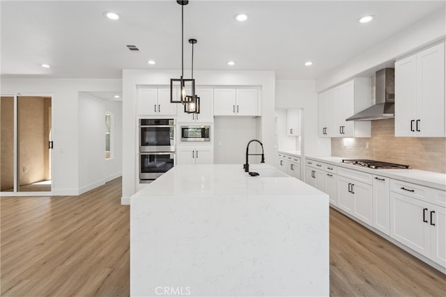 kitchen featuring light wood-type flooring, wall chimney range hood, hanging light fixtures, and a kitchen island with sink