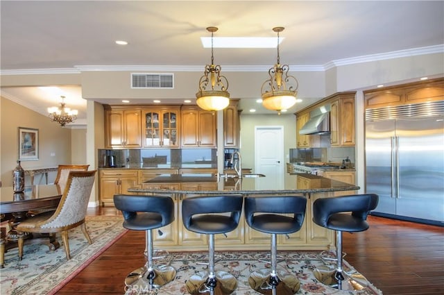 kitchen featuring wall chimney range hood, sink, a center island with sink, decorative backsplash, and stainless steel built in fridge