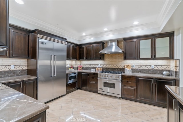 kitchen featuring light stone countertops, built in appliances, crown molding, dark brown cabinets, and wall chimney range hood