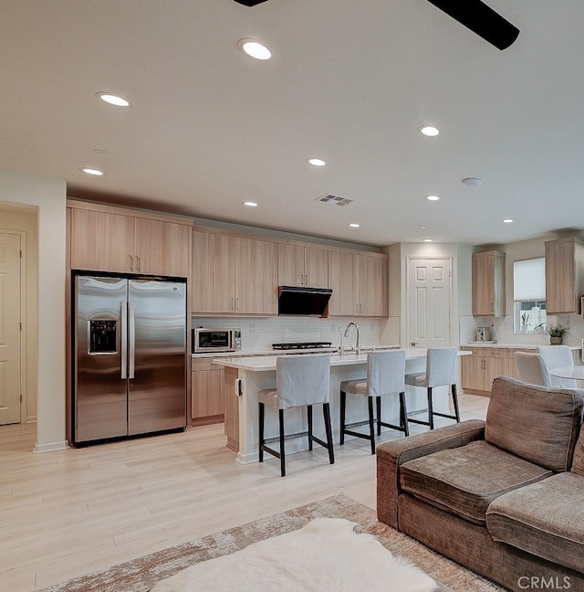 kitchen featuring exhaust hood, a center island with sink, appliances with stainless steel finishes, light brown cabinetry, and light hardwood / wood-style flooring