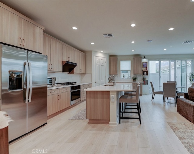 kitchen with light brown cabinetry, appliances with stainless steel finishes, light wood-type flooring, and an island with sink