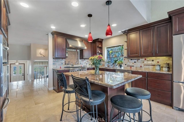 kitchen with stainless steel appliances, wall chimney range hood, light stone counters, pendant lighting, and decorative backsplash