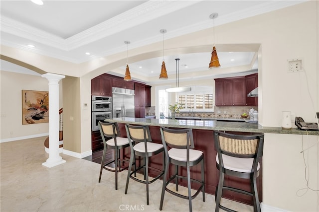 kitchen featuring a raised ceiling, hanging light fixtures, kitchen peninsula, stainless steel appliances, and decorative columns