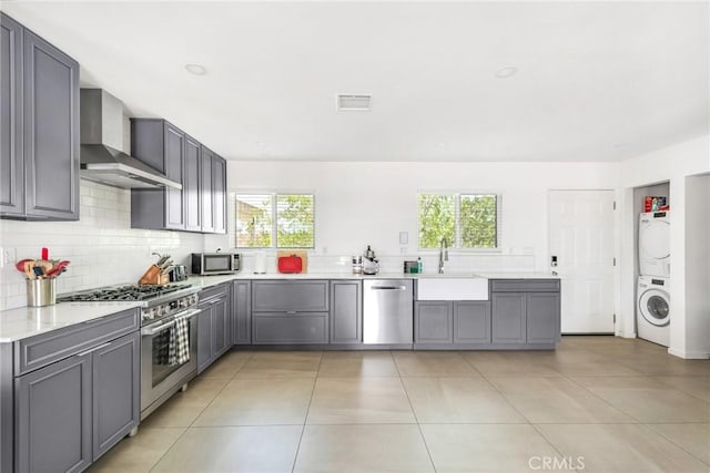 kitchen featuring stainless steel appliances, a sink, wall chimney exhaust hood, gray cabinetry, and stacked washer and dryer
