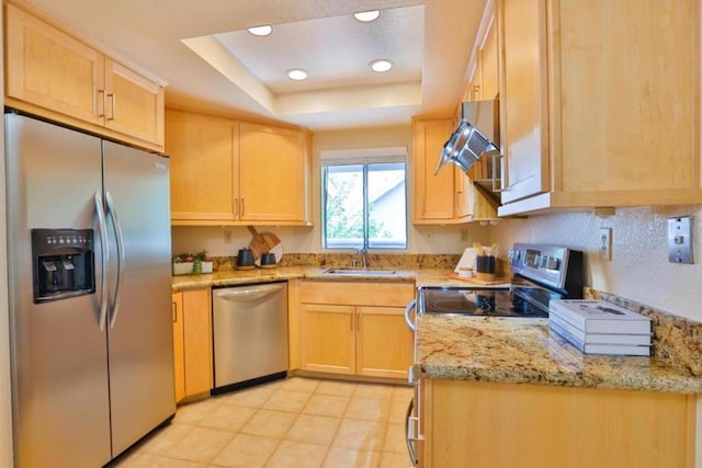 kitchen with stainless steel appliances, light brown cabinetry, sink, light stone counters, and a tray ceiling