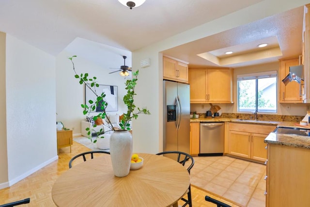 kitchen with light brown cabinets, ceiling fan, appliances with stainless steel finishes, light stone countertops, and a tray ceiling