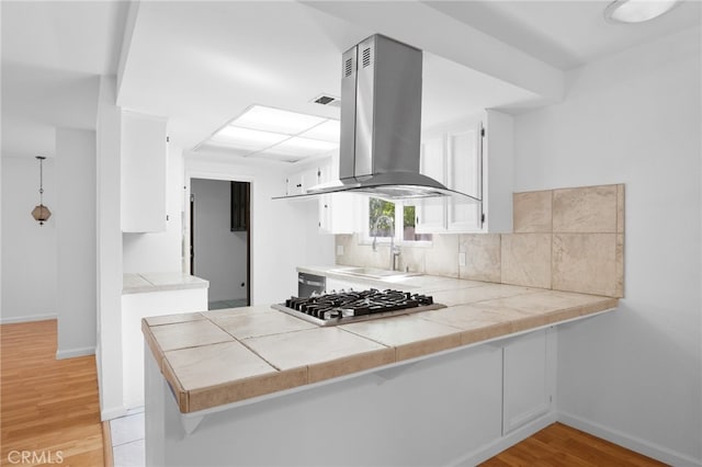 kitchen with tile counters, backsplash, light hardwood / wood-style flooring, wall chimney exhaust hood, and kitchen peninsula