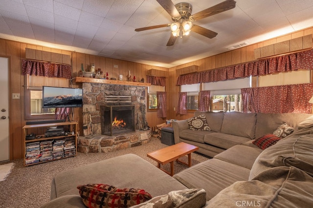 carpeted living room featuring ceiling fan, wooden walls, and a stone fireplace