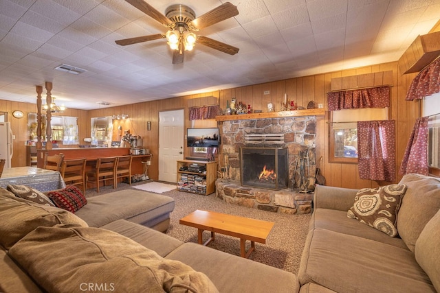 living room featuring ceiling fan, carpet flooring, wooden walls, and a stone fireplace