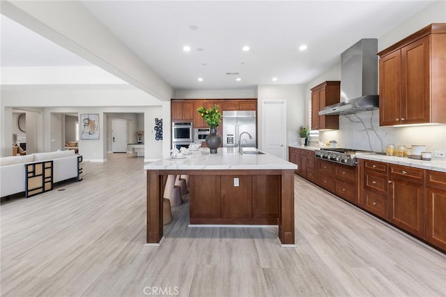 kitchen featuring appliances with stainless steel finishes, light wood-type flooring, sink, wall chimney range hood, and a center island with sink