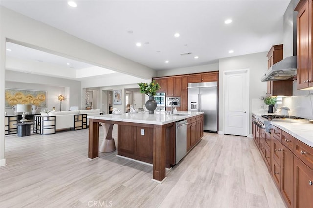 kitchen featuring a kitchen island with sink, sink, wall chimney exhaust hood, appliances with stainless steel finishes, and light hardwood / wood-style floors